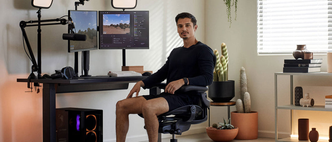 Gamer wearing all black sits in an Aeron Gaming Chair in front of his airy gaming set-up surrounded by monitors, a microphone and potted plants.