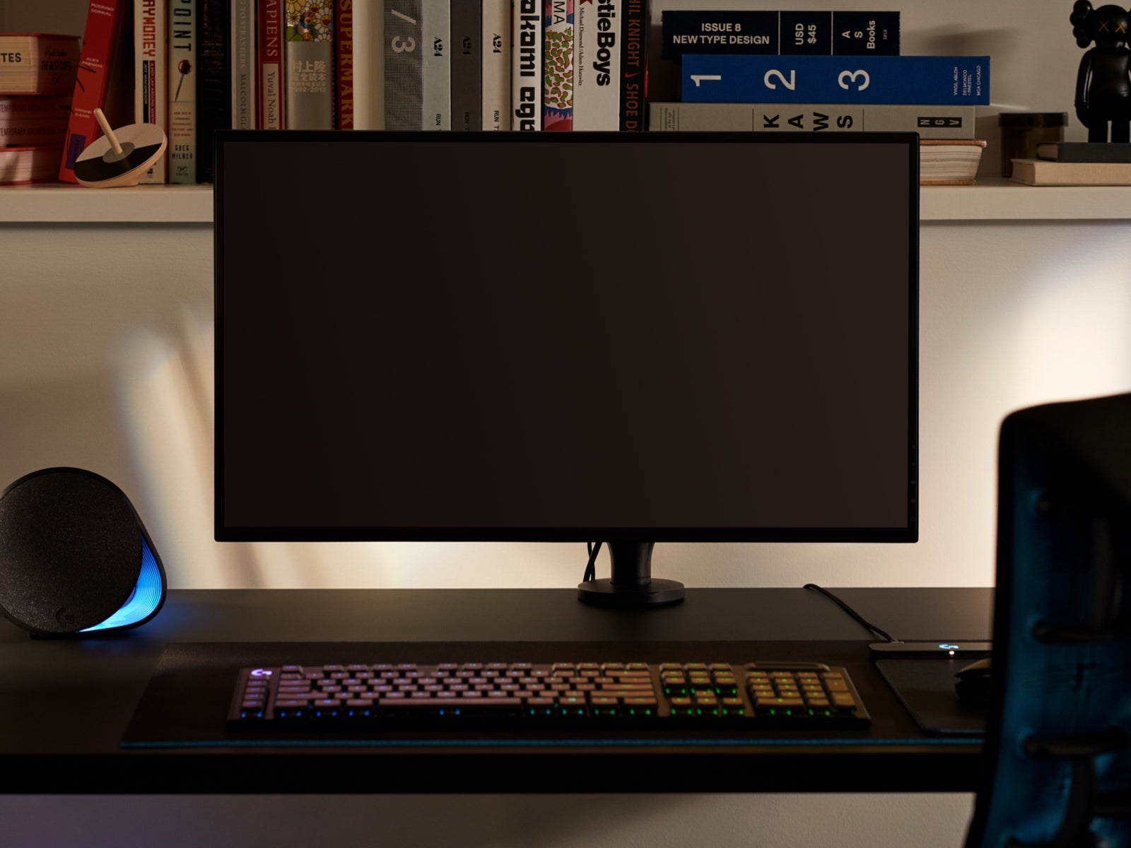A large monitor, representing the fact that the Ollin Monitor Arm can accommodate screens of all sizes, is attached to the arm on top of a desk with a keyboard in the foreground and items on a shelf in the background.
